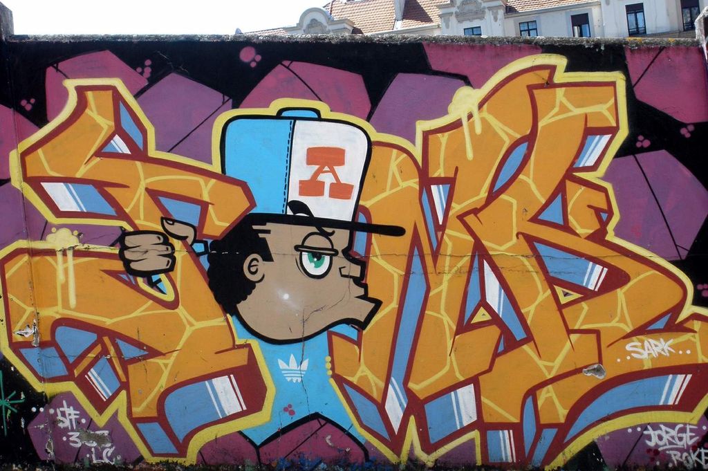 Fargerik graffiti på en mur. En person med kaps bryter seg vei ut mellom bokstaver i et ord. Foto.