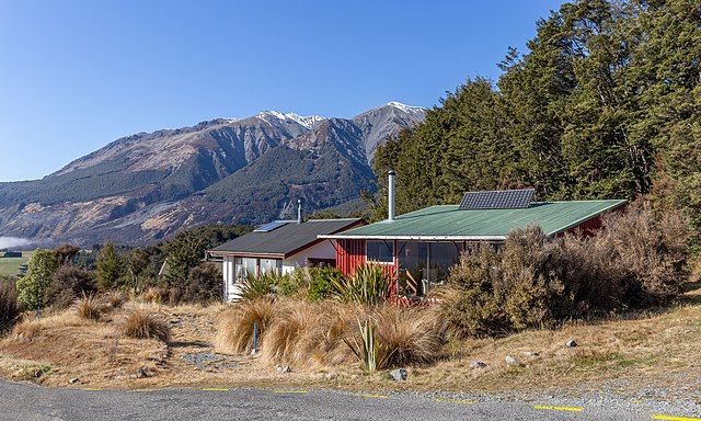 Photo: Two small houses, one red, one white. There are solar panels on the roof. In the background we see snow capped mountains and some water, like a fjord or lake. 