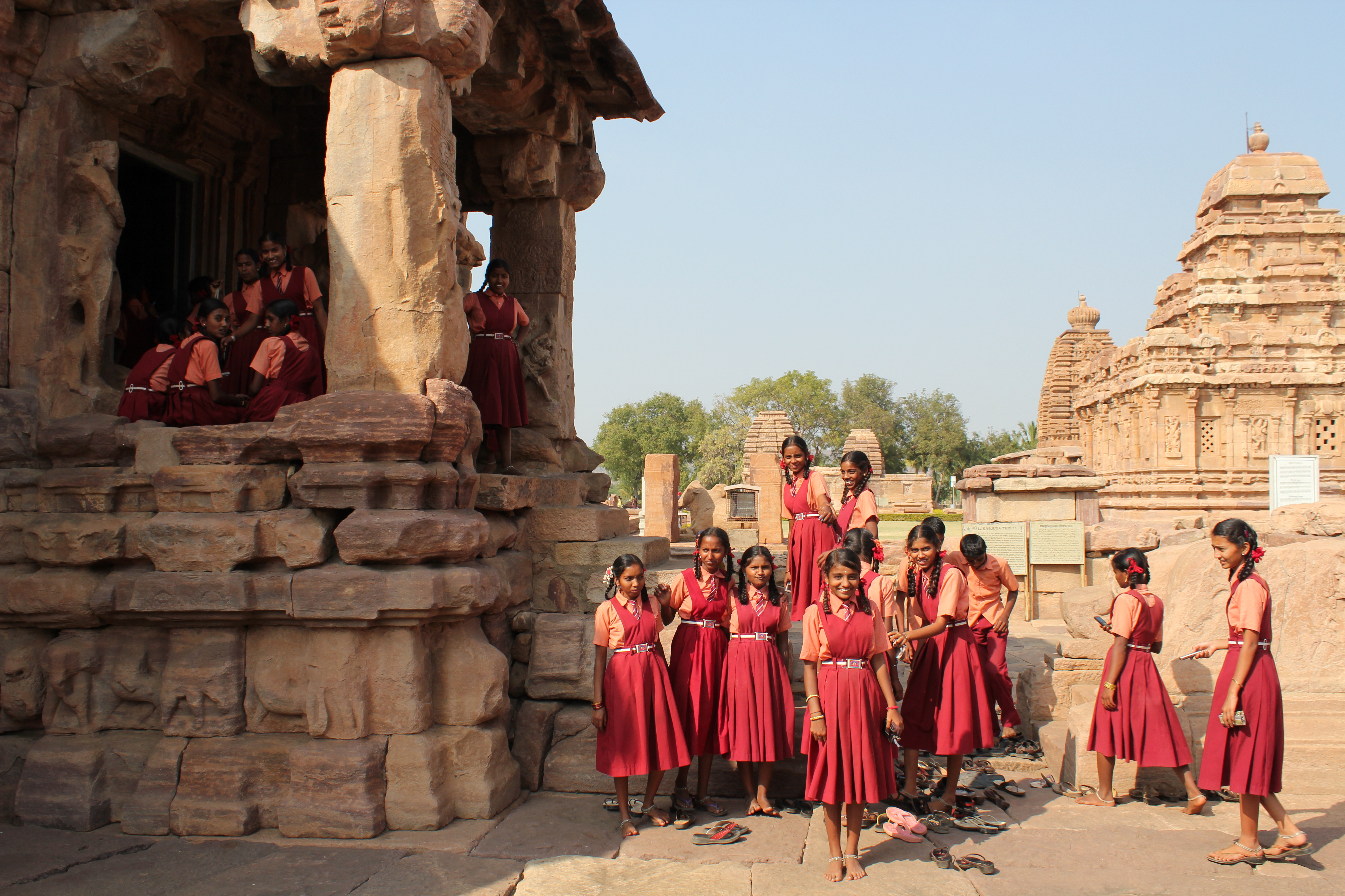 Two groups of Indian girls dressed in orange shirts and red dresses. They are posing in front of what looks like an ancient  temple. Photo.