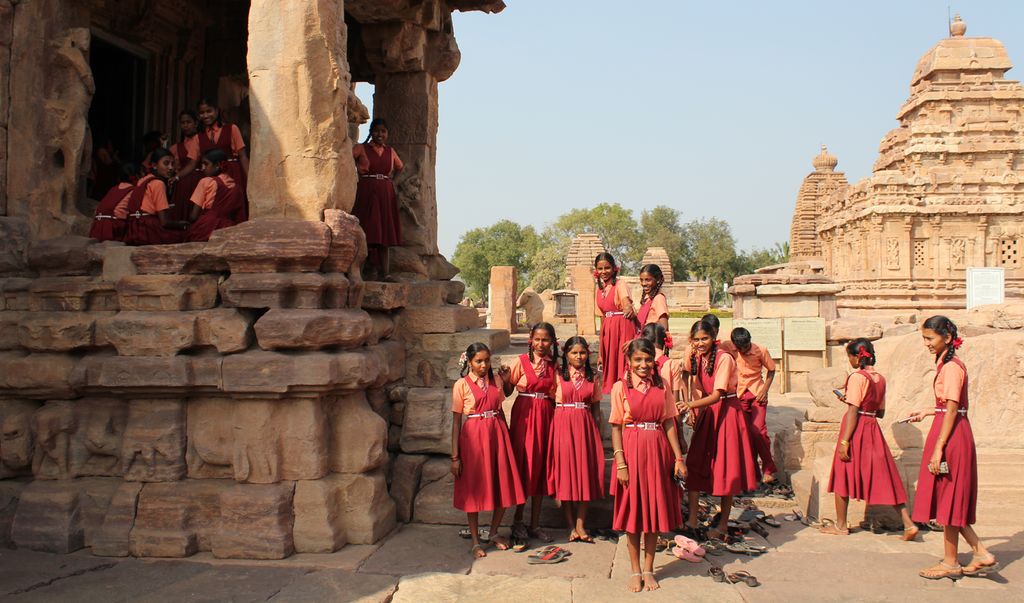 Two groups of Indian girls dressed in orange shirts and red dresses. They are posing in front of what looks like an ancient temple. Photo.