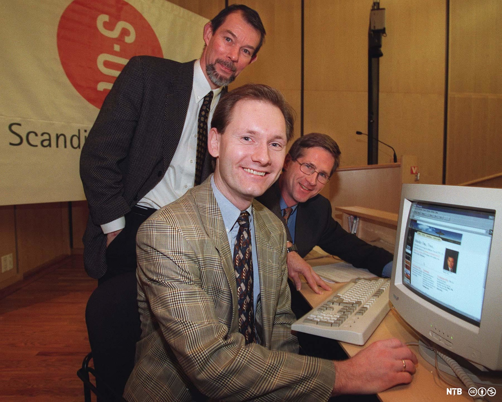 Tre menn foran en gammeldags datamaskin med bilderørskjerm. Foto.