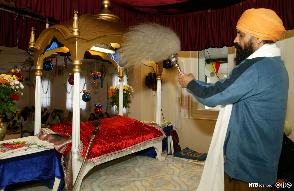 Mann med turban utfører et rituale foran en bok som ligger under et rødt klede og en baldakin. Foto.