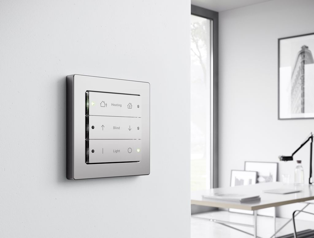 En vegg med påmonterte brytere med symboler og LED-indikatorer. Bryterne kan styre oppvarming, persienner og belysning. I bakgrunnen en pult og en kontorstol. Foto.