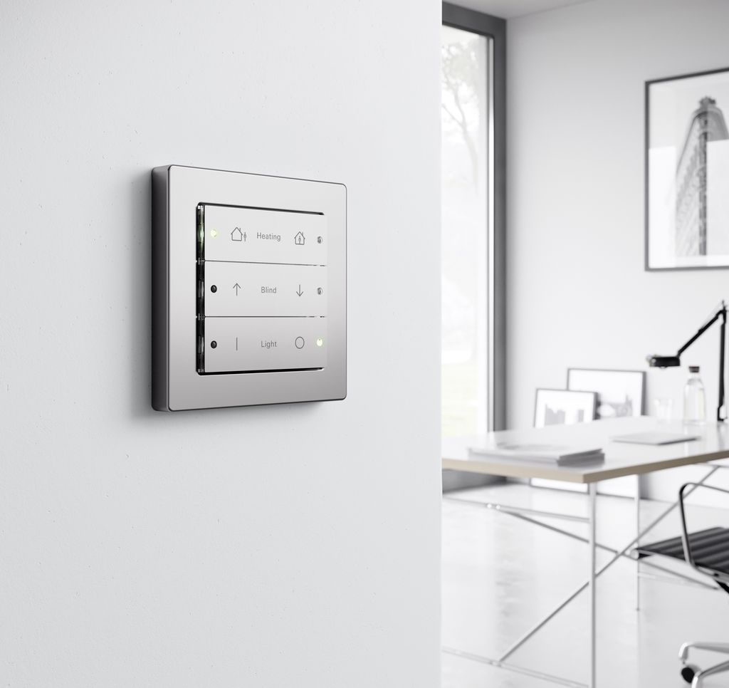 Vi ser ein kvit vegg med brytarar med symbol og LED-indikatorar. Brytarane kan styre oppvarming, persienner og lys. I bakgrunnen ser vi ein pult og ein kontorstol. Foto.