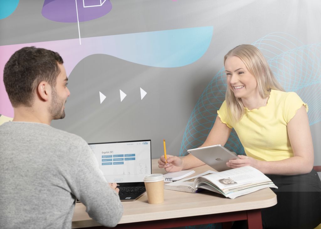 Gutt og jente arbeider sammen med pc, nettbrett og lærebok mellom seg på bordet. Foto.