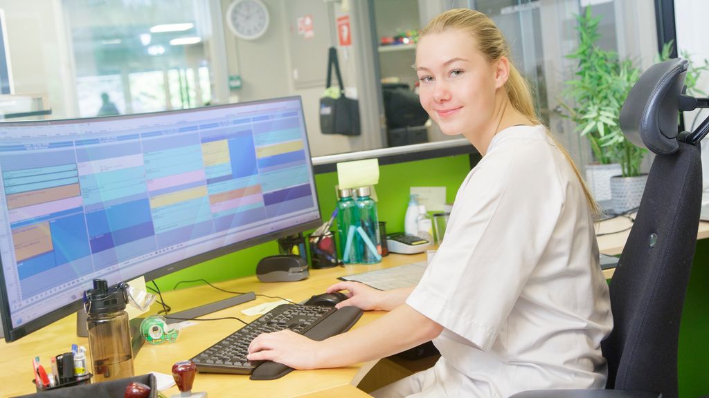 Helseservicearbeidar sit framfor pc i resepsjonen. Foto.