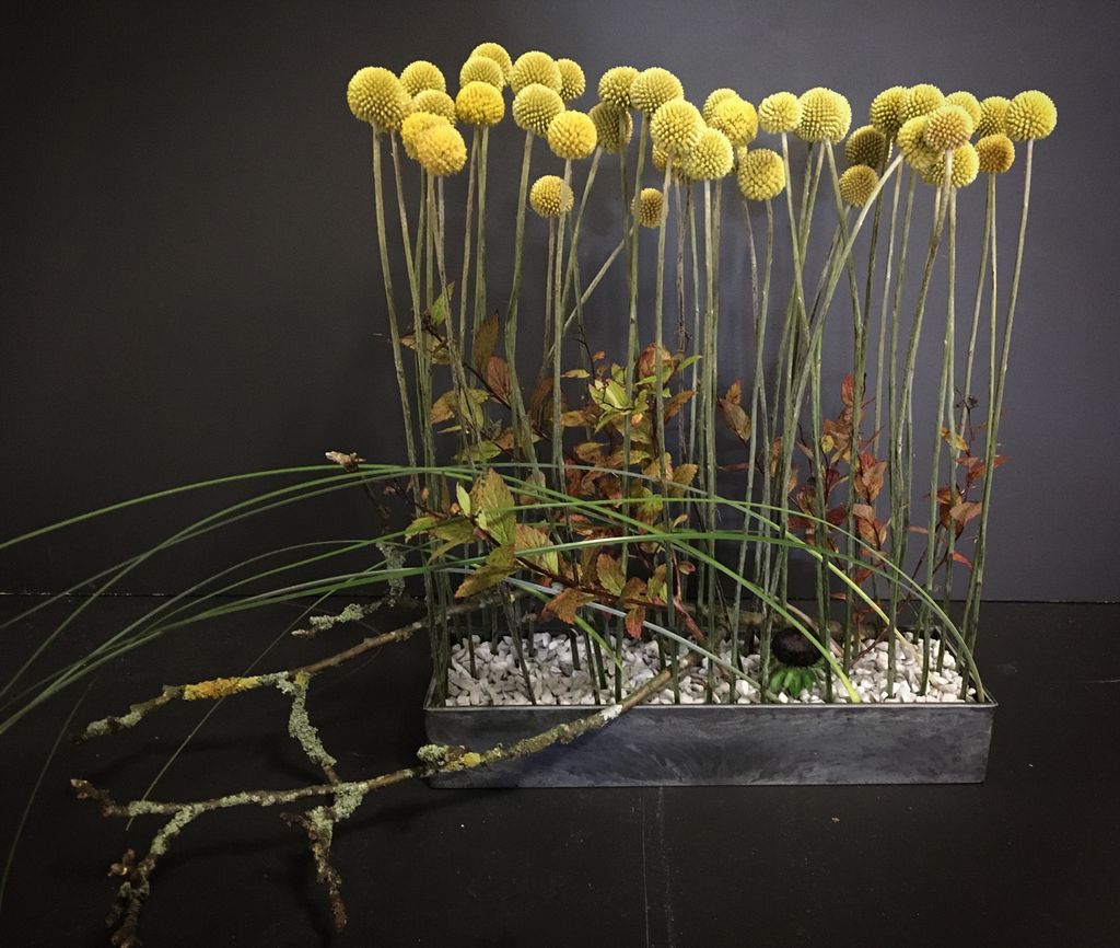 Blomsterdekorasjon med gule blomster, kvister og stein på et rektangulært brett. Foto.