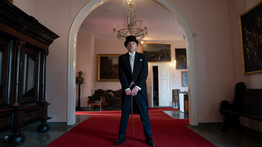 En mann på rundt femti år står midt i et stort rom. Det er høyt under taket. Langsmed veggene rundt ham står det tunge, gamle møbler, og på veggene henger det oljemalerier. Mannen er kledd i en staselig mørk dress med hvit skjorte og sløyfe. På hodet har han en svart flosshatt. Han støtter seg på en spaserstokk og ser alvorlig og bestemt inn i kamera. Foto.