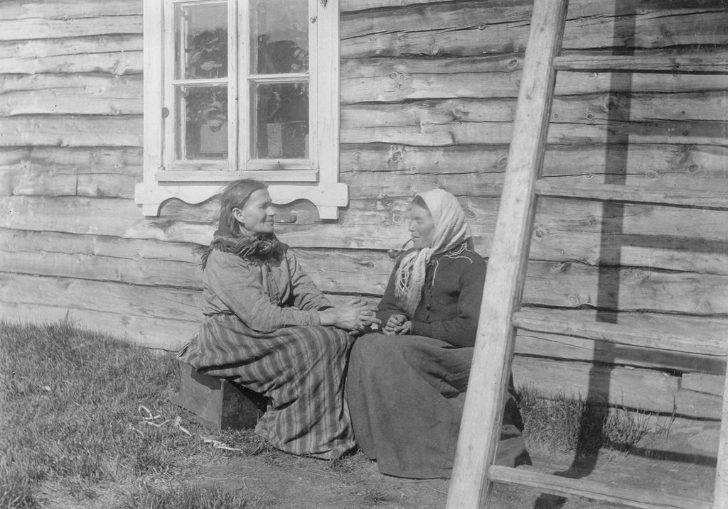 To kvinner i langt skjørt, ei med sjal rundt halsen og ei med skaut, sitter utenfor et trehus. Begge kvinnene røyker pipe. Svart-hvitt foto.