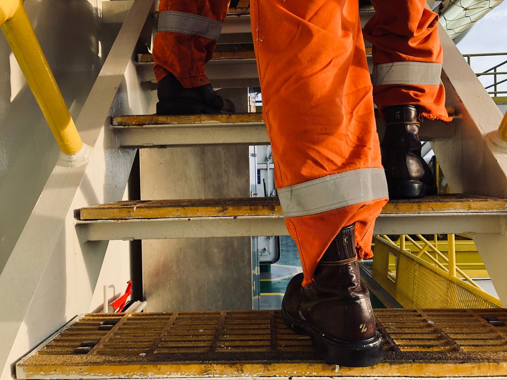 Føtter på vei opp i ei trapp offshore. Personen har på seg vernesko og kjeledress. Foto.