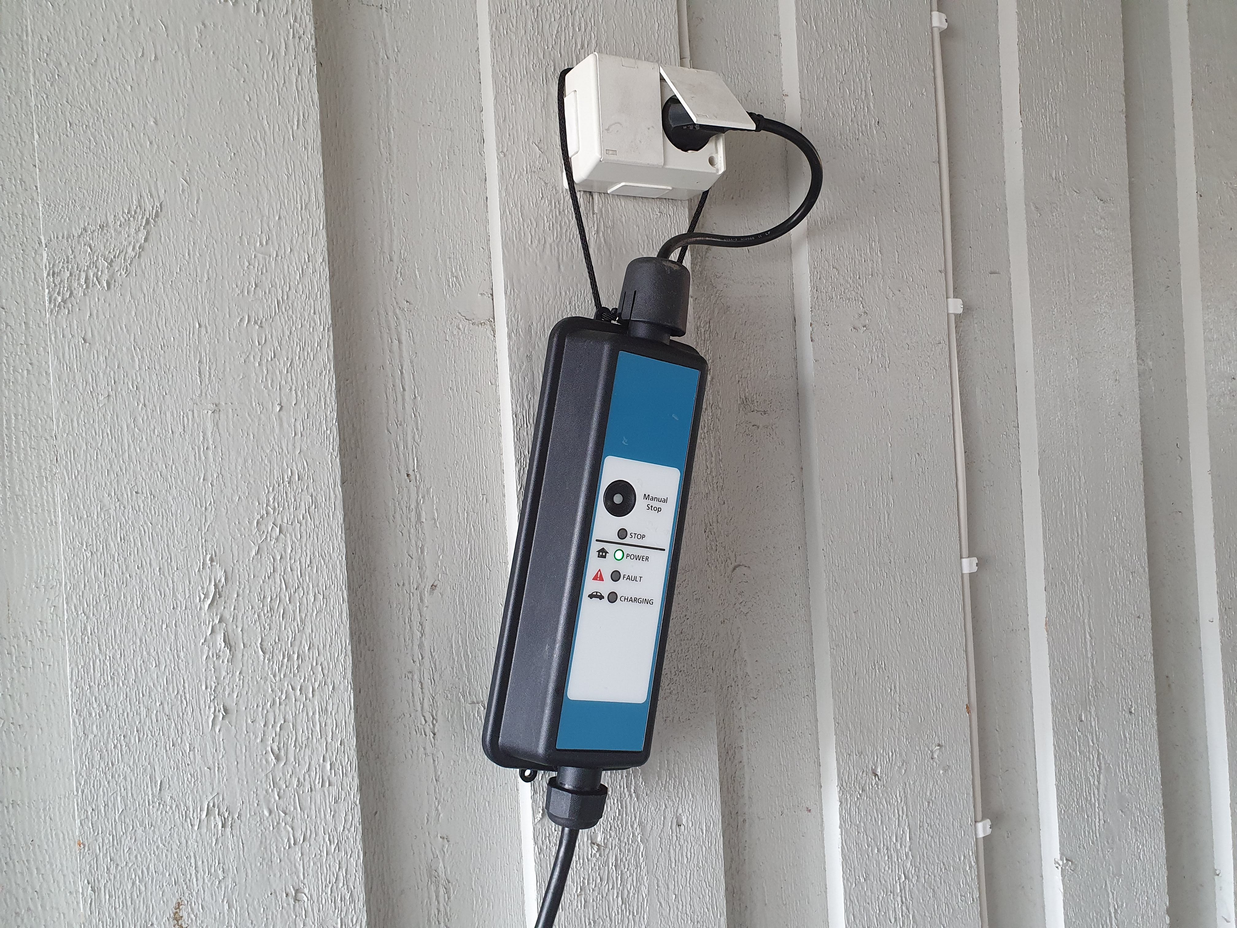 Ladeboks for elbil, som er plugget inn i en vanlig utendørs stikkontakt montert på en husvegg av tre. Ladeboksen henger i stikkontakten. Foto.