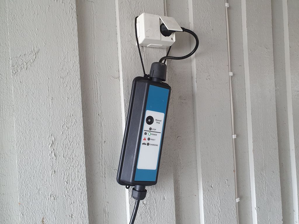 En ladeboks for elbil er plugget inn i en vanlig utendørs stikkontakt montert på en husvegg av tre. Ladeboksen henger i stikkontakten. Foto.