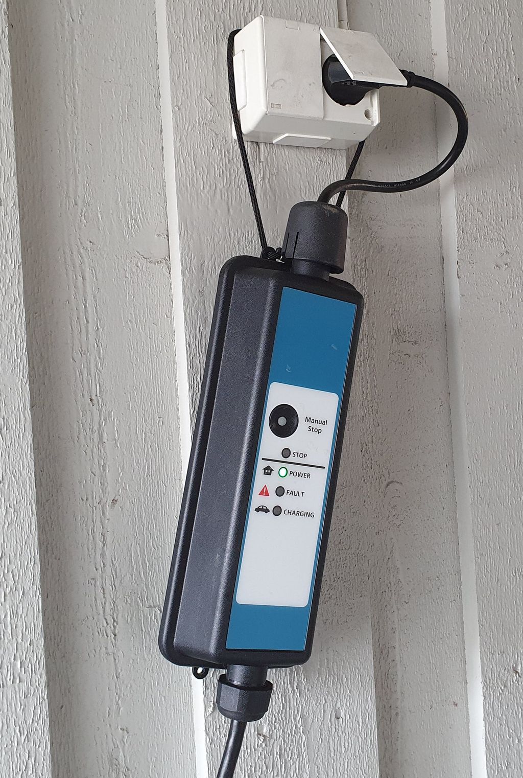 Ladeboks for elbil, som er plugget inn i en vanlig utendørs stikkontakt montert på en husvegg av tre. Ladeboksen henger i stikkontakten. Foto.