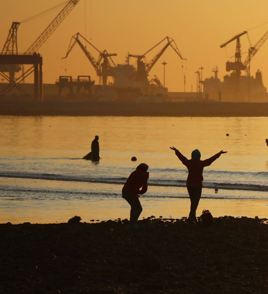 Silhuetter av folk på strand foran industriområde i solnedgang. Foto.
