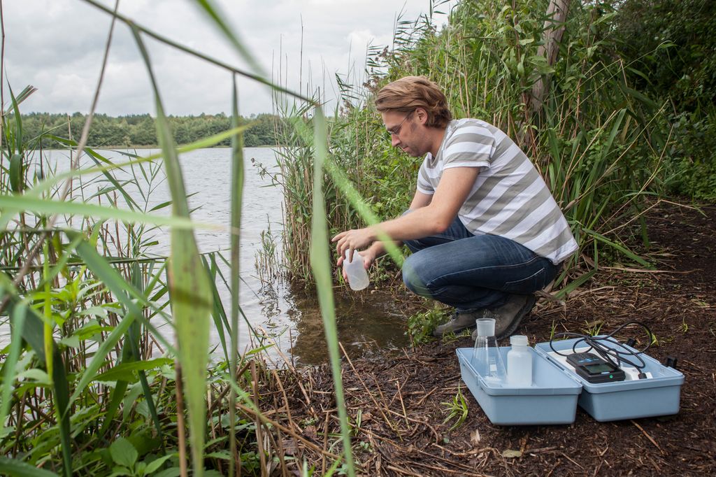 Mann tar vannprøve fra innsjø. Ved siden av seg har han en boks med diverse prøveflasker og et måleinstrument. Foto.
