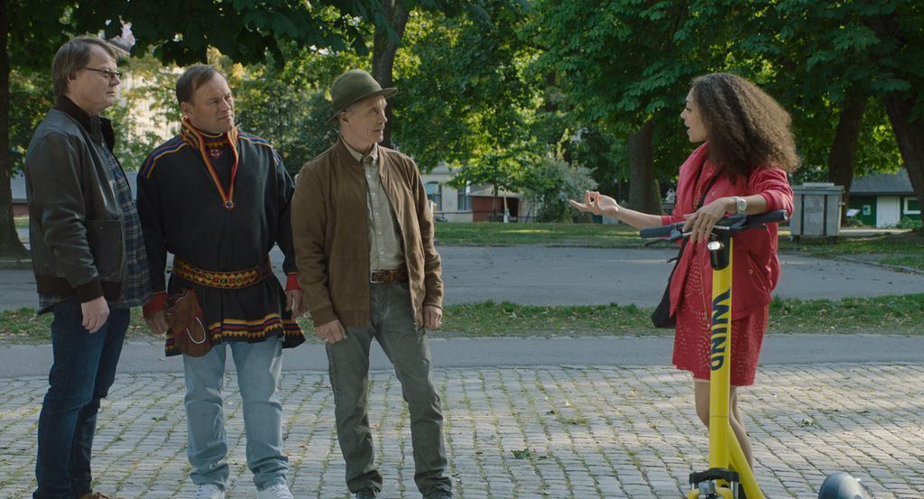 Utsnitt fra filmen "Koftepolitiet": Tre menn, to av dem kledd i ordinære klær og den midterste i samekofte, snakker med en urban dame med rød kjole, langt, mørkt hår og gul, elektrisk sparkesykkel. Foto.