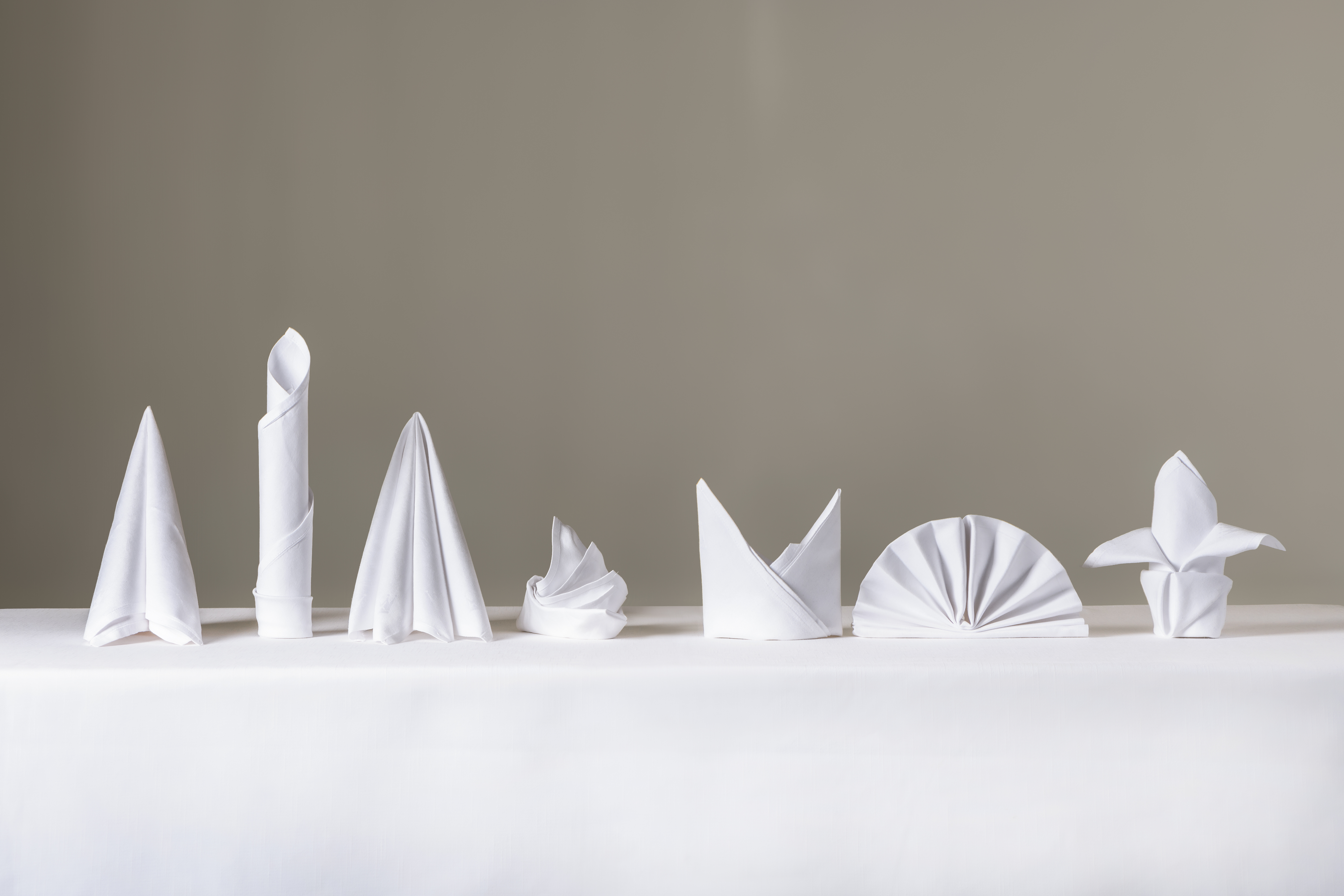 Sju kvite tøyserviettar er bretta på ulike måtar og står på eit bord med kvit duk. Foto