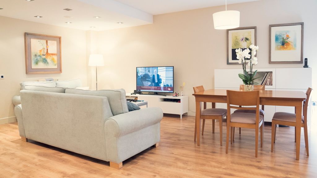 Ei stue med to lyse sofaer, tv og et bord med fire stoler rundt. Den ene sofaen er vendt mot tv-en, det er en orkidé på bordet og malerier på veggene. Foto.