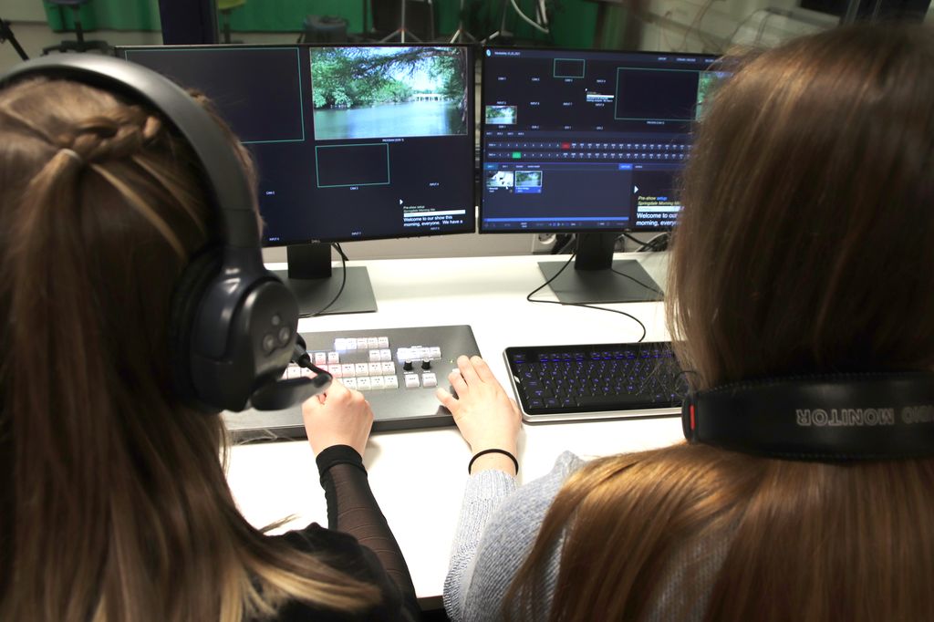 To jenter med hodesett ser på skjermer i et TV-kontrollrom. De har hendene på en bildemikser, og bildet er tatt mellom dem bakfra. Foto. 