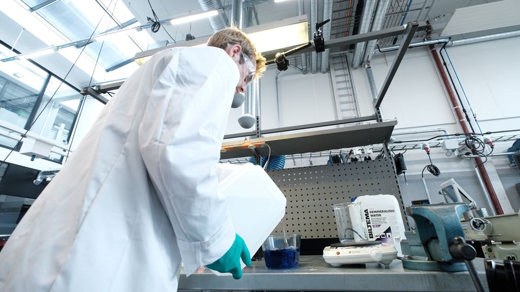 Person i hvit frakk med vernebriller og beskyttelseshansker heller kjemikaler fra en plastdunk til en beholder på en arbeidsbenk. Foto.