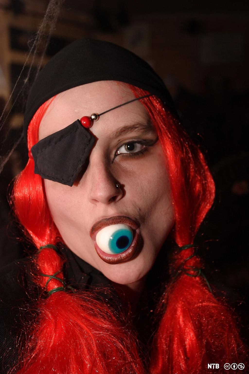 Ung kvinne med rød parykk med musefletter, svart tørkle på hodet, svart lapp over det ene øyet og et øyeeple i munnen. Foto.
