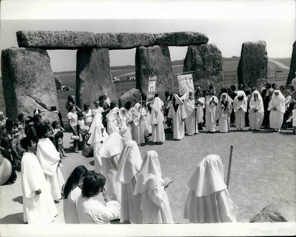 Mennesker kledd i hvite kapper står i en stor ring ved Stonehenge i England. Svart-hvitt. Foto.