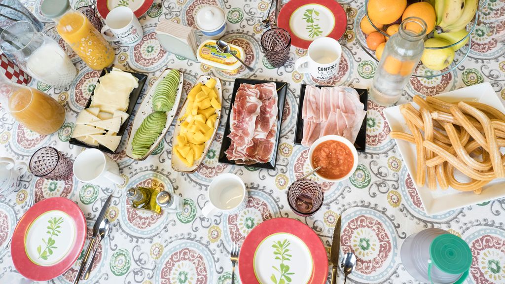 Fugleperspektiv av bord med mønstret duk og ulike spanske tapasretter på fat: to ulike hvite oster på samme fat, oppskåret avokado og mango, oppskåret spekeskinke, oppskåret kokt skinke, churros. Flasker med eplejus, melk, appelsinjus og vann. En flaske med flytende sjokolade. En skål med hakkede tomater. En bolle med bananer, epler og appelsiner. Liten flaske med olivenolje. Saltkar. Sukkerskål. Et beger med smør og en skje oppå lokket. Det er dekket på med asjett, kniv, gaffel, skje, glass og kopp til fire personer. Foto.