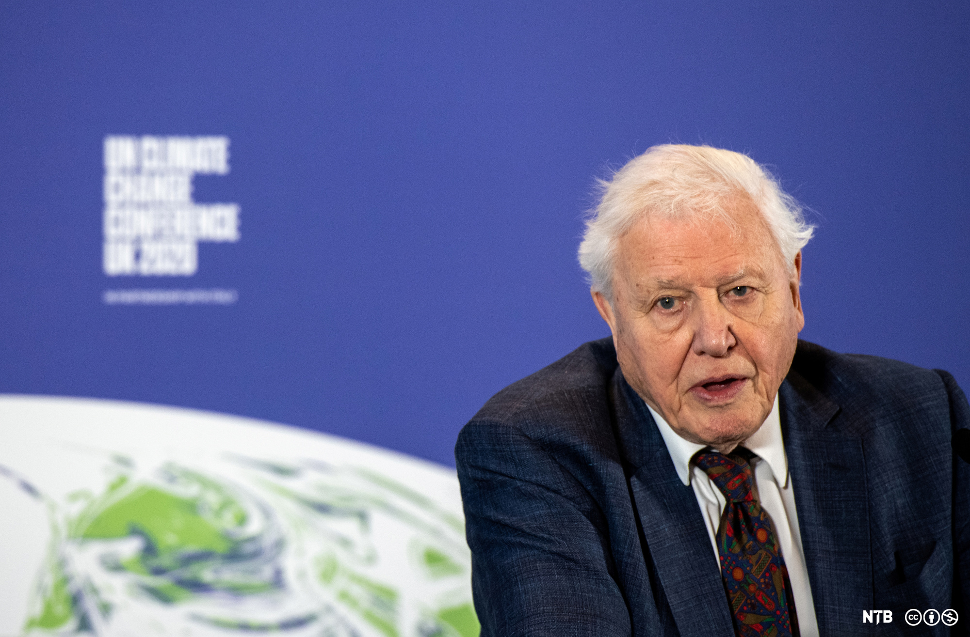 David Attenborough i mørk dress. Han er plassert framfor eit bilete av jorda og logoen til klimakonferansen. Foto.