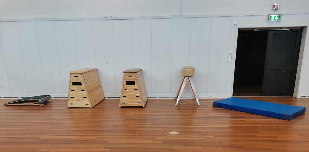 Springbrett, to kasser, en bukk og ei matte i en gymsal med hvit vegg. Foto.