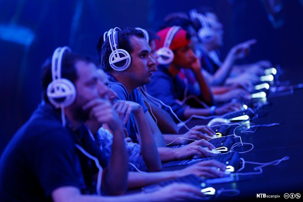 Flere menn med hodetelefoner sitter på rekke med hvert sitt tastatur og hver sin datamus. Foto.