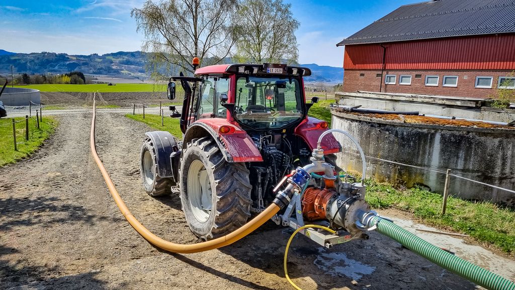 En traktor står ved et fjøs. Det er koplet ei gjødselpumpe til traktoren, og en slange strekker seg nedover en liten vei og langt ut på et grønt jorde hvor det er en annen traktor langt unna. Foto.