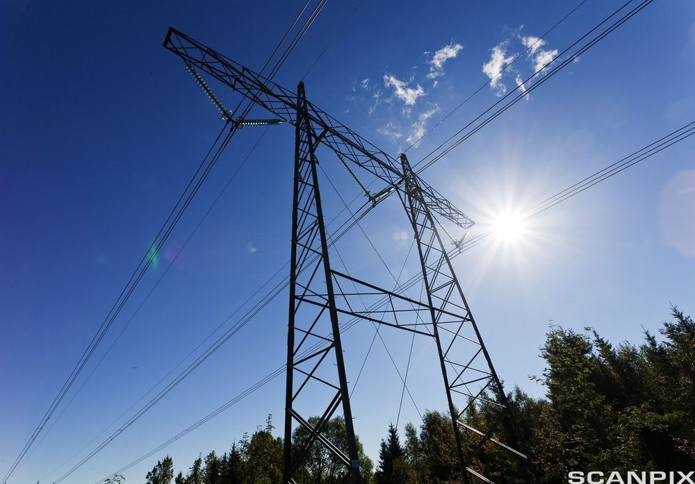 Kraftmast med høyspent. I bakgrunnen ser vi bartrær, blå himmel og sol. Foto. 