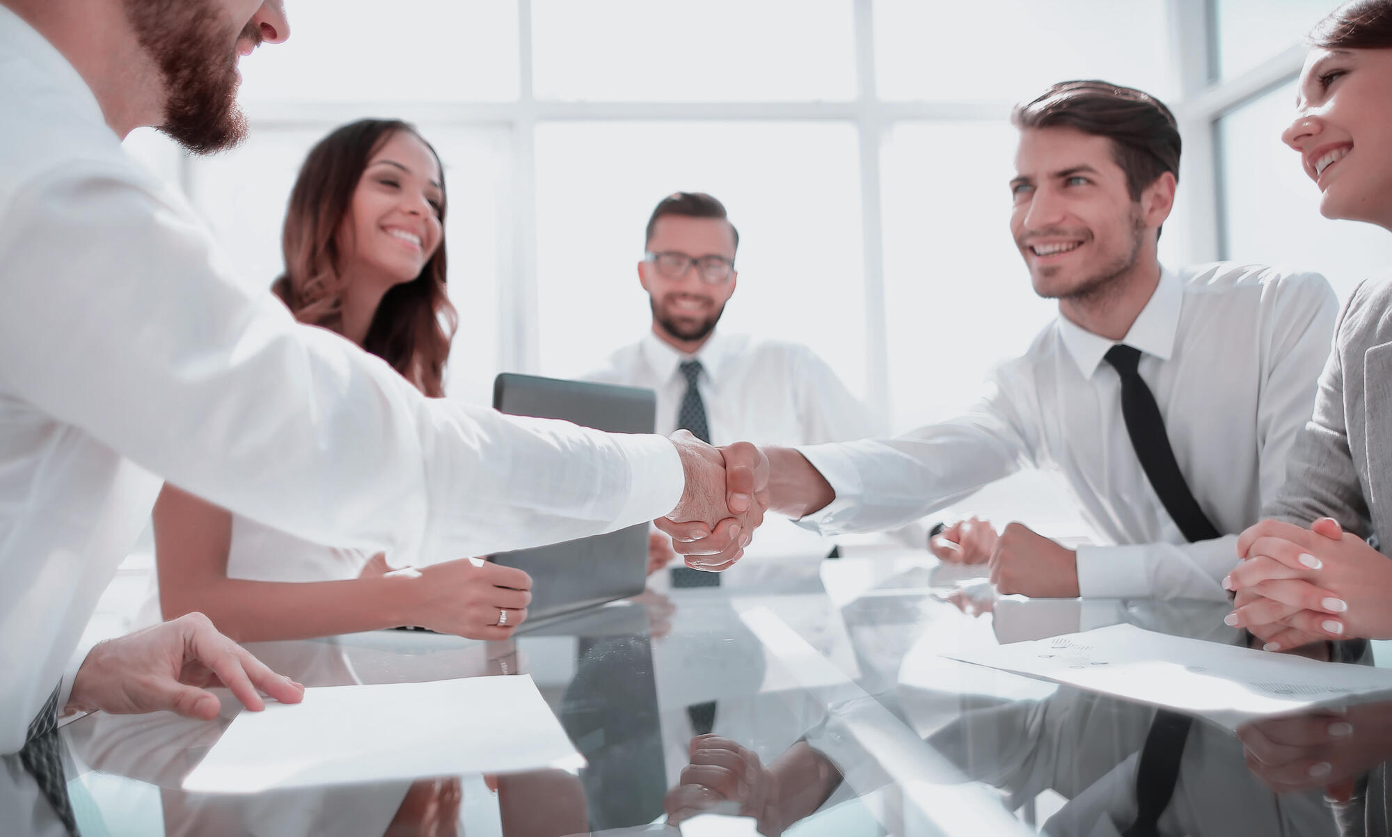 Fire personer i formelt antrekk rundt et bord. De smiler. To av dem tar hverandre i hånden over bordet. Foto.