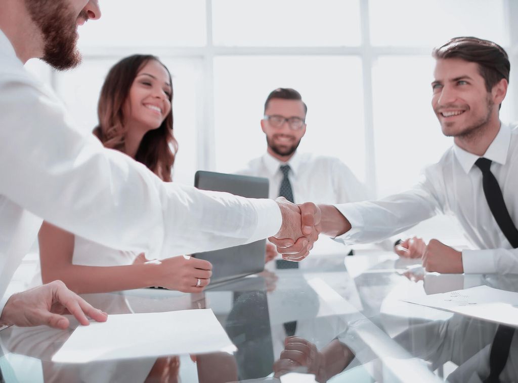 Fem personar i formelt antrekk rundt eit bord. Dei smiler. To av dei tek kvarandre i handa over bordet. Foto.