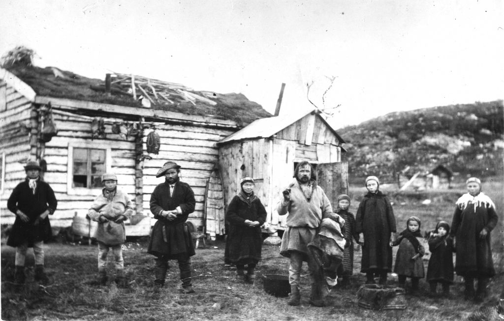 Ei gruppe sjøsamer fotografert på slutten av 1800-tallet. Her er både menn, kvinner og barn i kofter foran et trehus. Foto. 