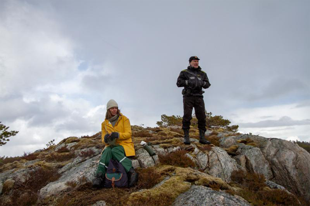 En kvinne i gul regnjakke og grønn regnbukse sitter på en knaus. En politimann i uniform står litt lenger borte på knausen med hendene i lommene. Foto.