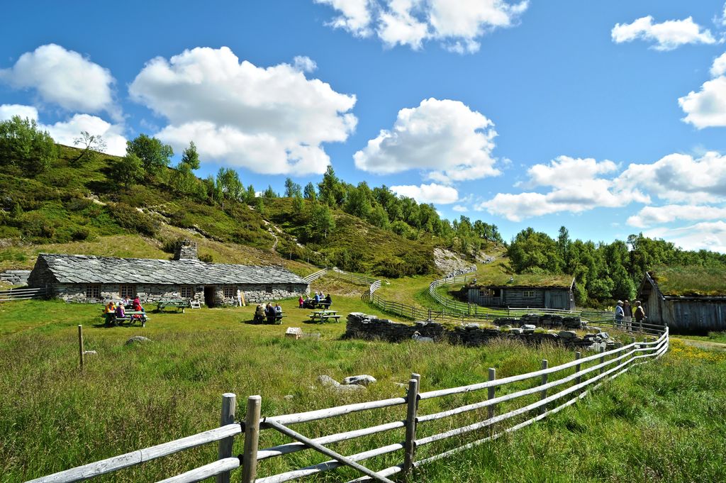 Landskap med flere lave seterbygninger. Det sitter mennesker på noen benker på grassletta foran det største huset. Dette huset er ganske langt og murt opp av naturstein og har skifertak. To andre, mindre hus er bygd av tømmer og har torvtak. Det er også et gjerde rundt setertunet. Foto.