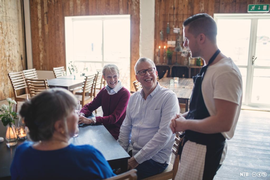 En ung mannlig servitør tar opp bestilling fra et bord med eldre gjester. Foto.