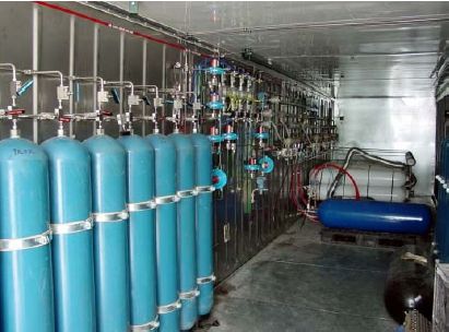 Sju lyseblå gassflasker, røyr og ventilar som står samankopla på ein vegg i eit rom. Foto.