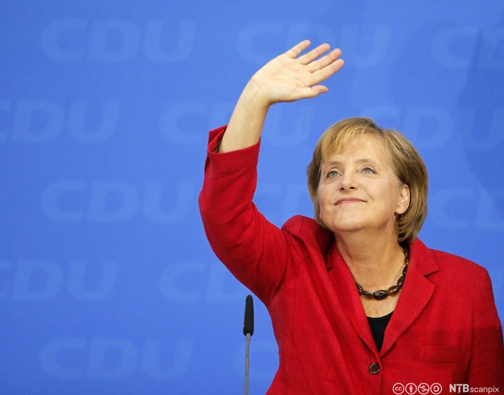 Den tyske politikeren Angela Merkel på et partimøte. Hun har på seg en rød blazer over en svart genser og et halskjede satt sammen av store, mørke, avlange steiner. Foto.