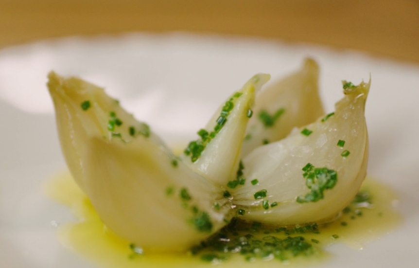 En bakt løk som er delt slik at den ligner en blomst, anrettet med gressløksmør på en hvit tallerken. Foto.