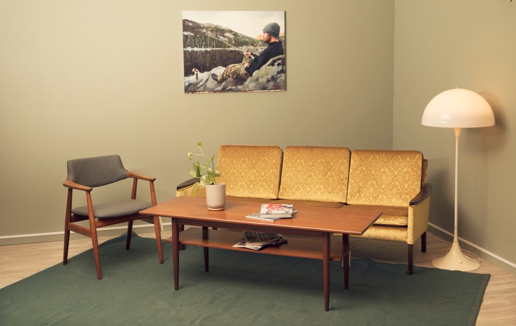 En grå stol, et teak-bord, en hvit stålampe og en gul sofa. Foto.