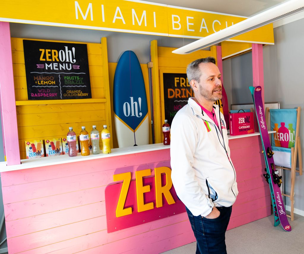 En person står foran en markedsføringsinstallasjon for et produkt. Installasjonen er utformet som en disk for kjøp eller servering og er preget av sterke ei gult og rosa. Et surfebrett, en strandstol og et par slalomski er brukt i installasjonen. Foran på disken står det ZERoh! Fire drikkeflasker er plassert oppå disken. Foto. 