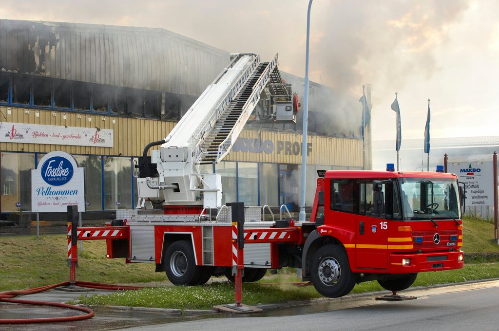 En redningsbil med kran står utenfor en stort Maxbo-bygg. Svart og grå røyk velter ut av vinduer i hele andre etasje i bygget. Foto.