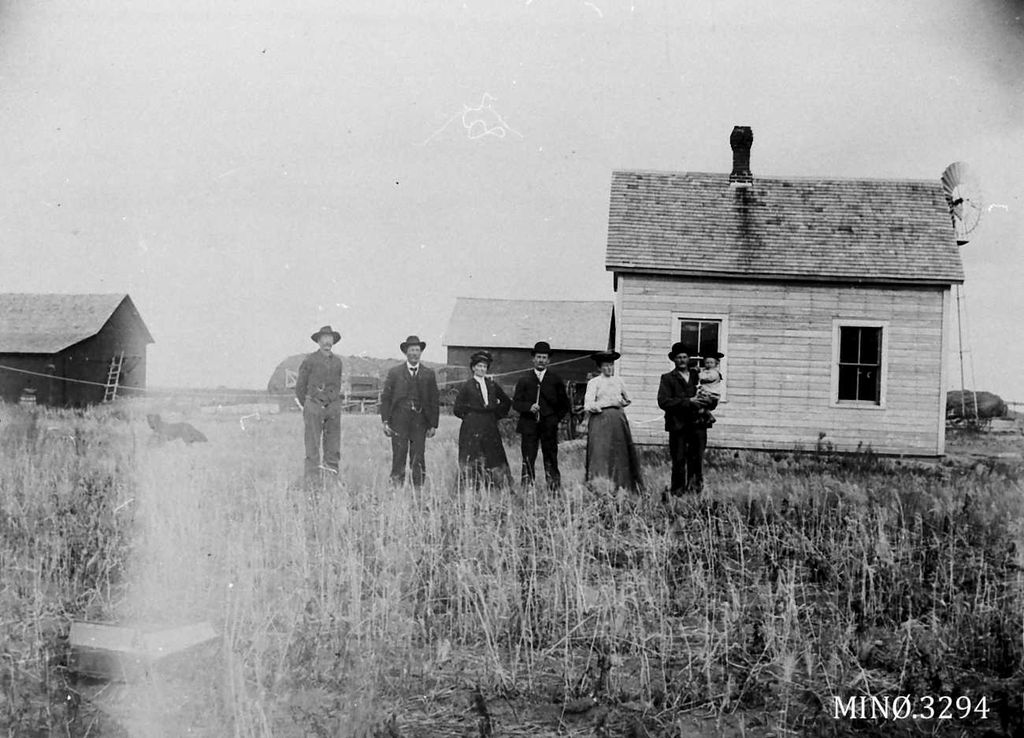 En familie står foran et nybygd hus med uthus.  Svart-hvitt-foto. 