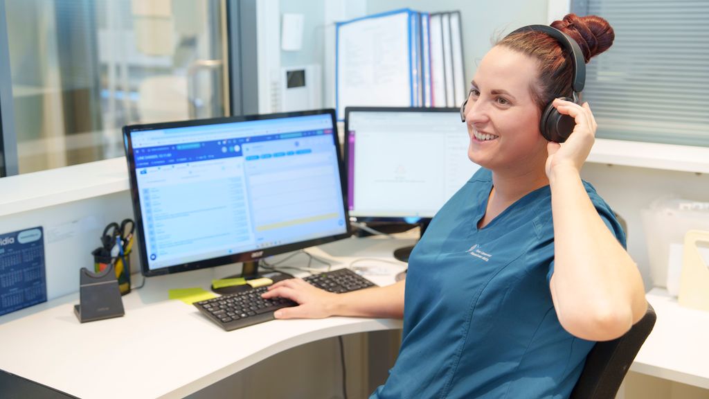 Helsesekretær sitter foran pc og justerer på hodetelefonene sine. Foto.