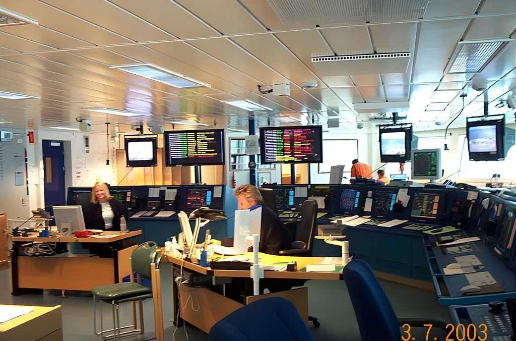 To personer sitter ved hver sin pult i et åpent kontorlandskap. Rundt dem er det flere kontrollbord med mange stasjoner, skjermer og kontrollpaneler. Kontrollrom på oljeplattform. Foto.