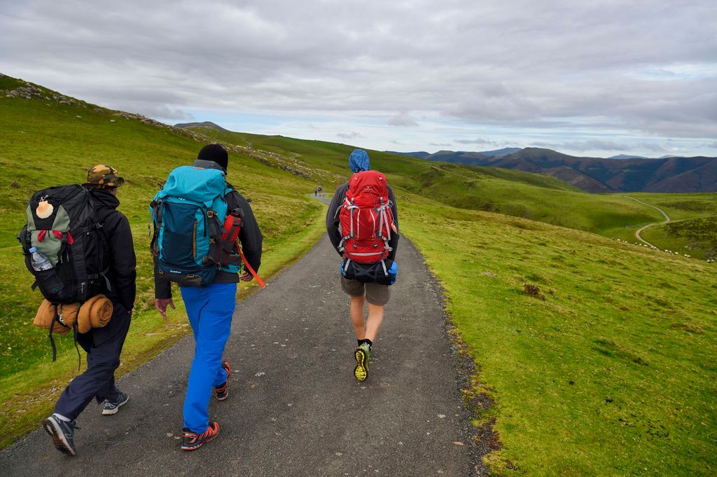 Tre personer med ryggsekker vandrer langs en sti. Det er et kupert landskap med grønne enger og fjell i bakgrunnen. Foto.