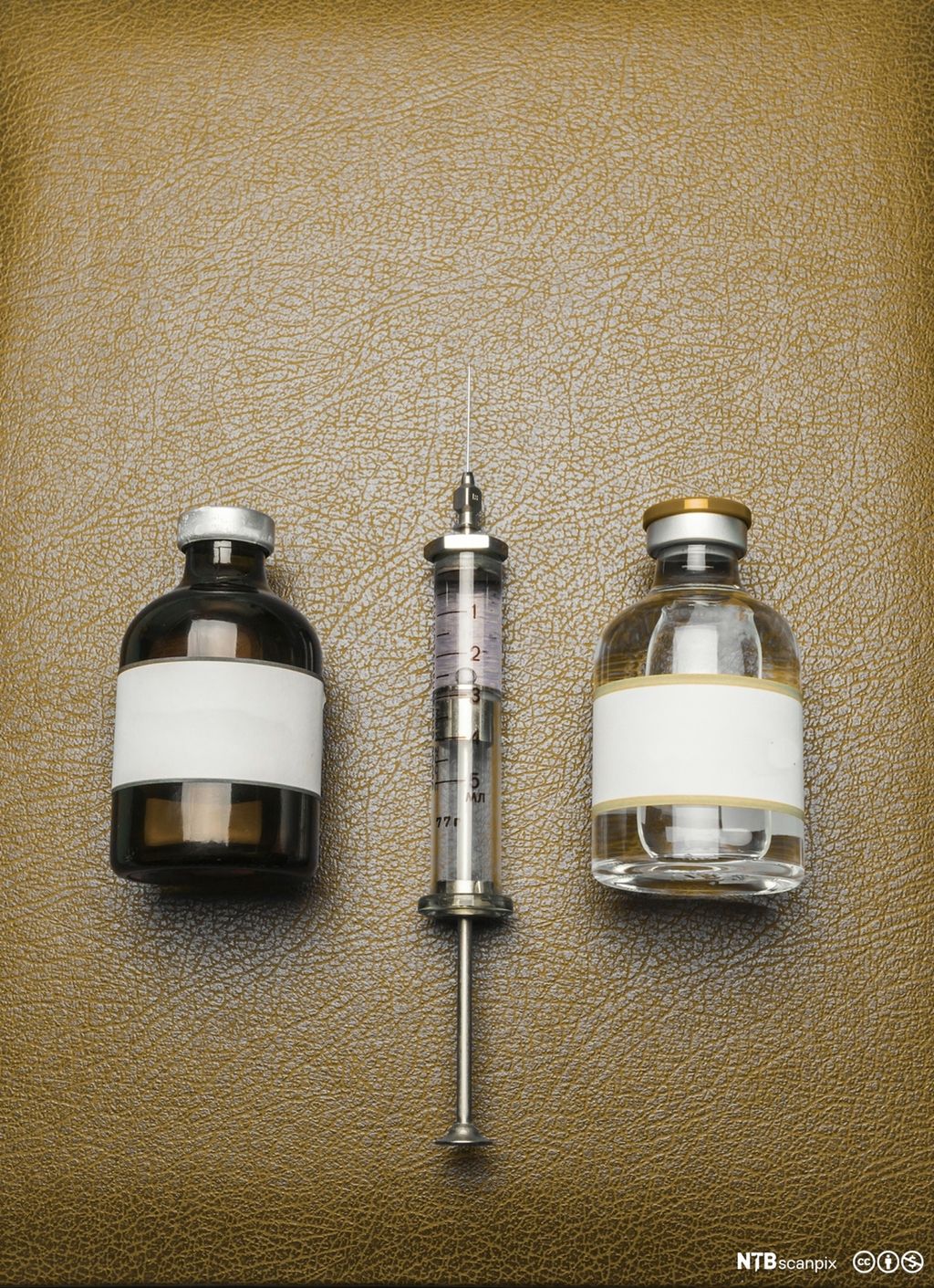 To medisinflasker i glass med skrukork og ei sprøyte. Sprøyta ligger i midten med ei medisinflaske på hver side. Foto.