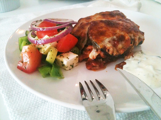Moussaka, et kjøttstykke med saus og en hvit dressing anrettet på en hvit tallerken. Foto.